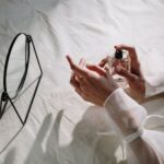 Delicate hands holding a perfume bottle beside a mirror on a white cloth background.